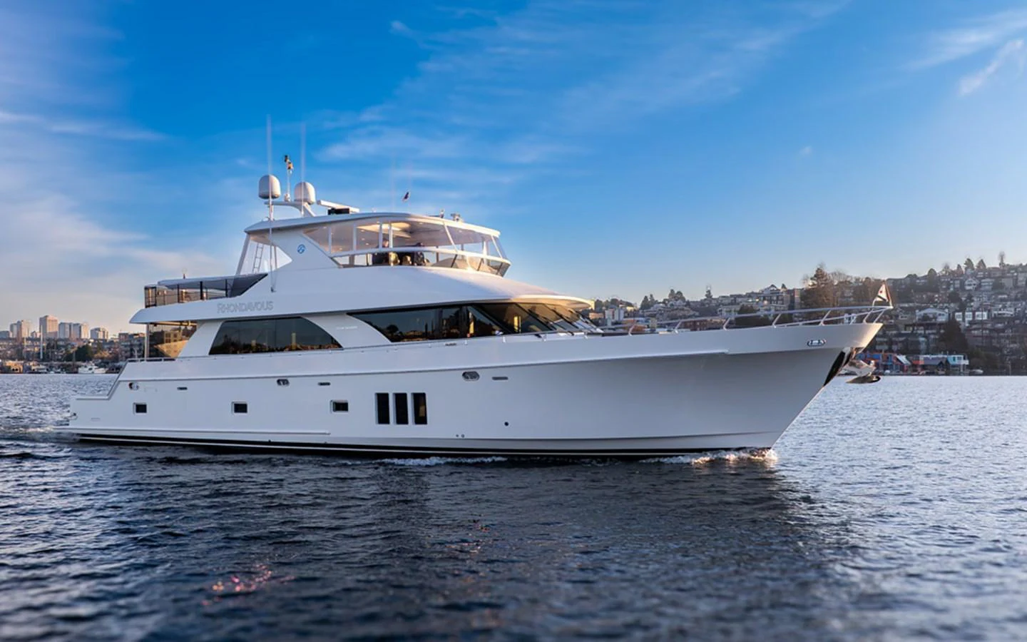 A sleek and modern yacht cruises gracefully on calm waters against a backdrop of a coastal cityscape. aboard RHONDAVOUS Yacht for Sale