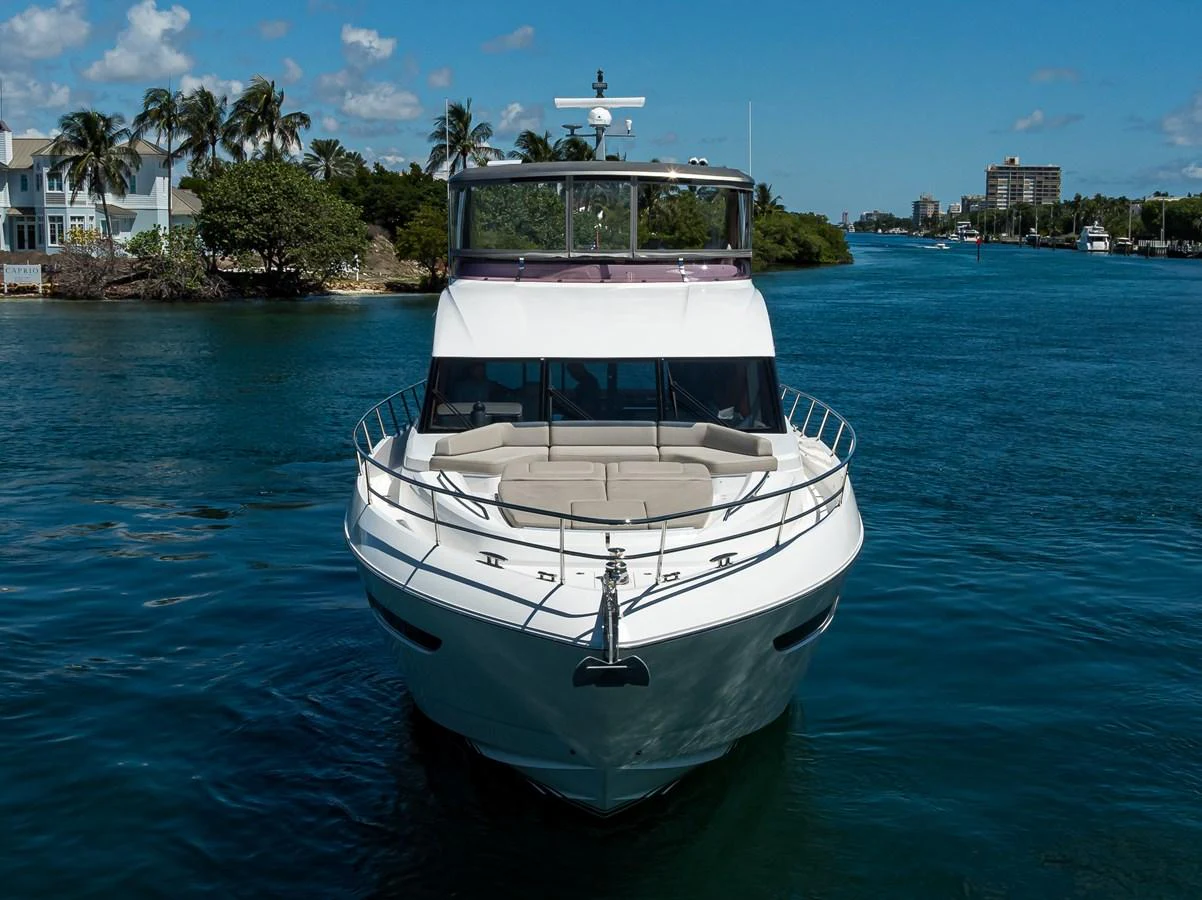 A sleek yacht is pictured head-on, cruising through a serene waterway with lush greenery and buildings in the background. aboard MISS AMY Yacht for Sale