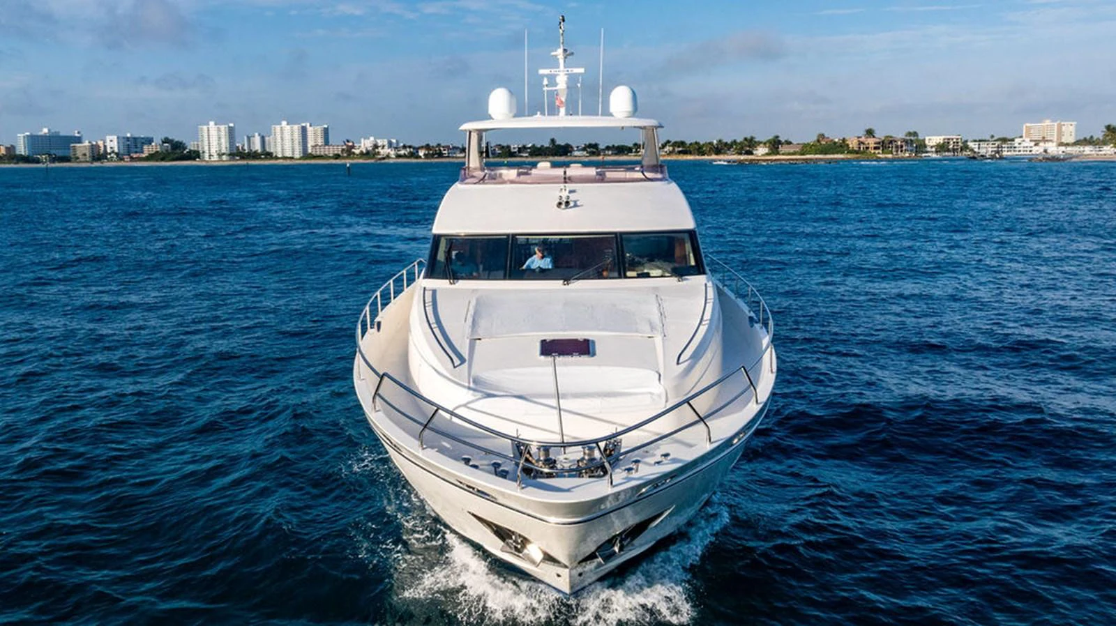 A luxurious yacht cruises forward on the open water with a cityscape in the background. aboard OASIS Yacht for Sale