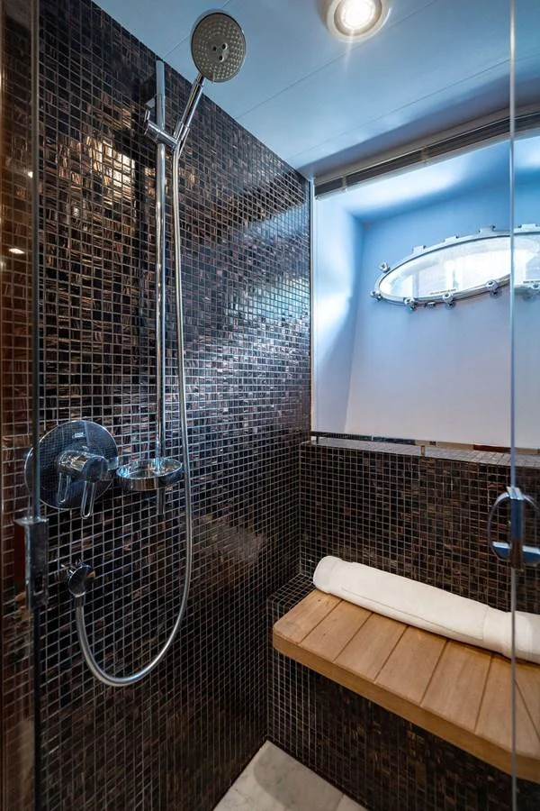 A modern yacht bathroom features a sleek shower area with dark mosaic tiles and a wooden bench. aboard OASIS Yacht for Sale