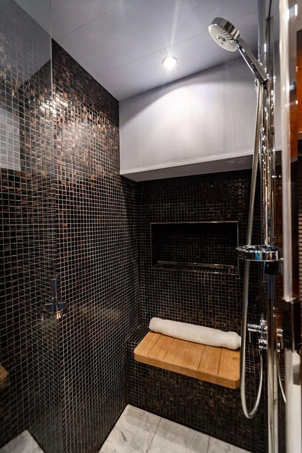 A luxurious yacht bathroom featuring a sleek shower area with dark mosaic tiles and a wooden bench. aboard OASIS Yacht for Sale
