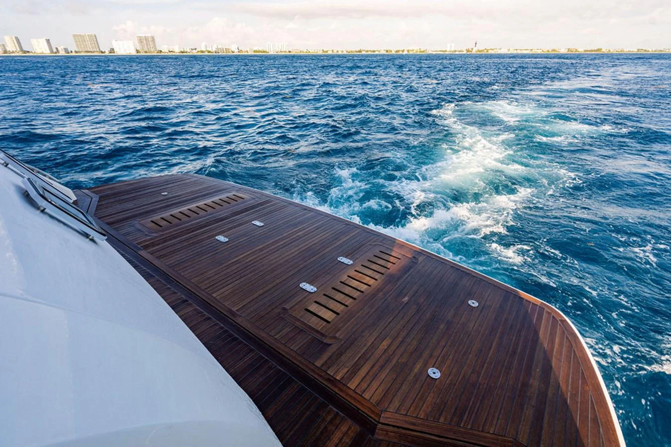 The image captures the elegant wooden aft deck of a yacht with a view of the open sea and distant shoreline. aboard OASIS Yacht for Sale