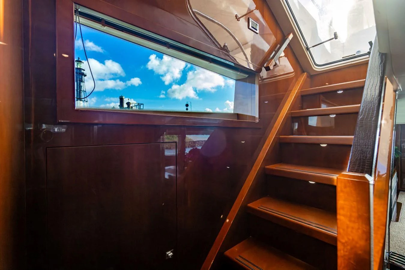 A polished wooden staircase leads to an upper deck, illuminated by natural light from a large window. aboard OASIS Yacht for Sale