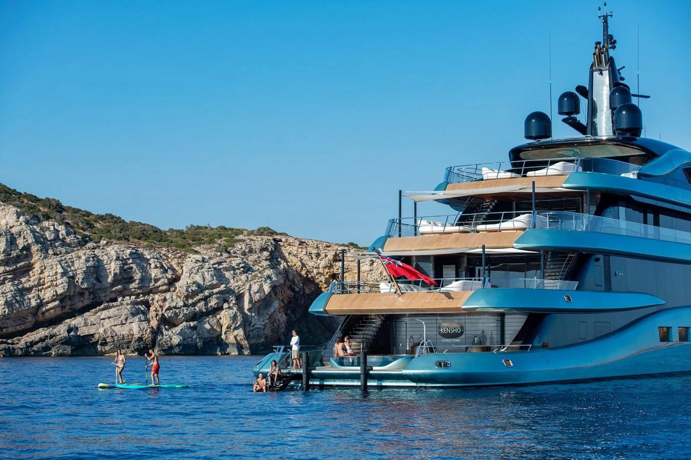 A luxurious yacht anchored near a rocky coastline with people enjoying water activities. aboard KENSHO Yacht for Sale