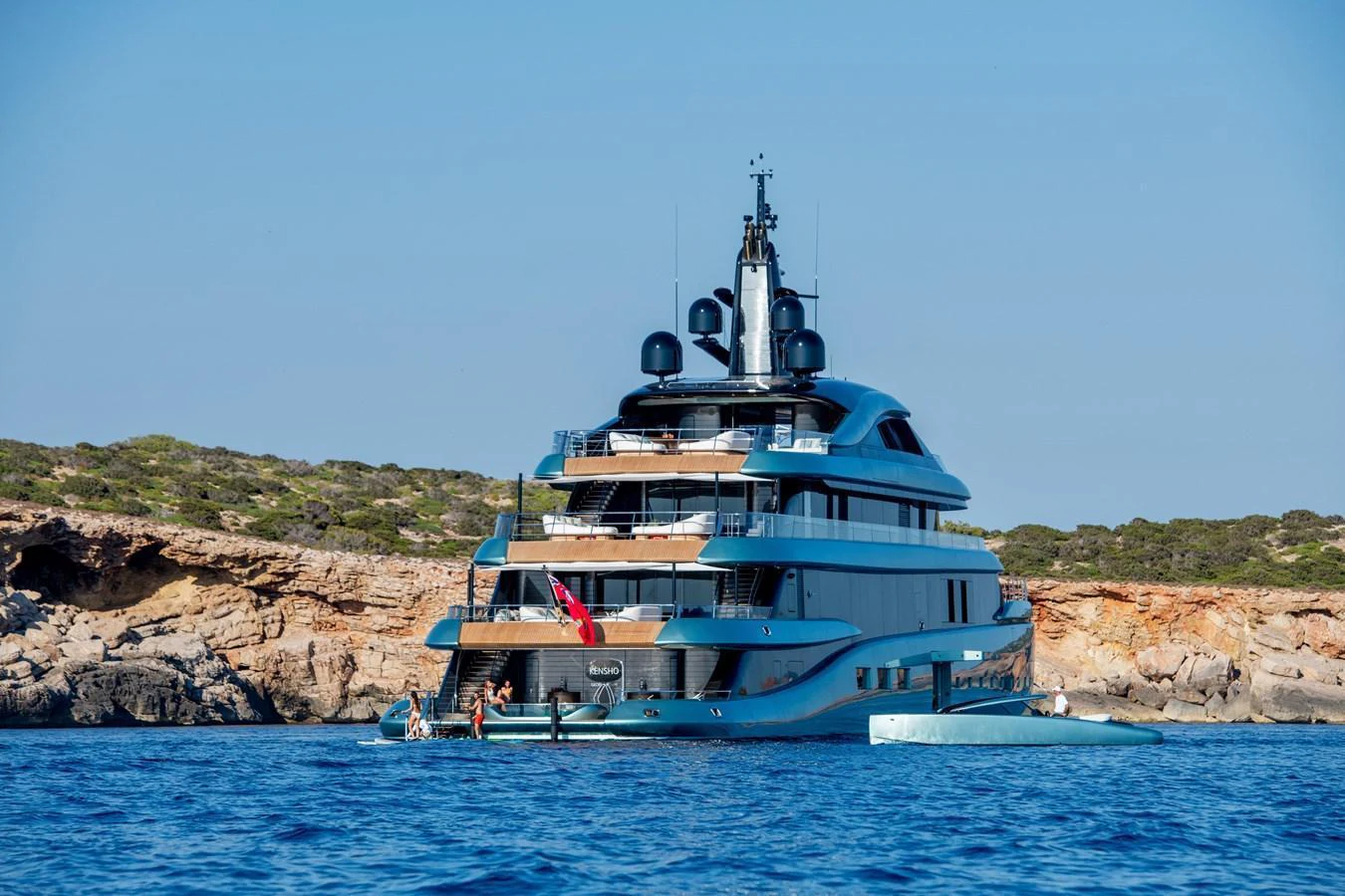 A luxurious yacht anchored near a rocky coastline with people enjoying water activities. aboard KENSHO Yacht for Sale