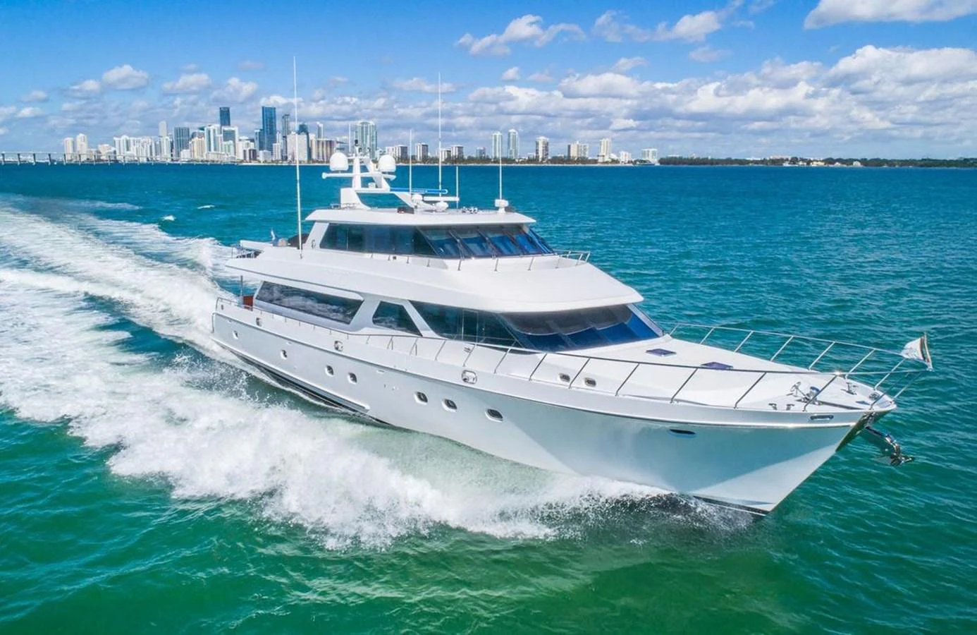 A sleek yacht cruises through vibrant blue waters with a city skyline in the background. aboard ROBIN'S NEST Yacht for Sale