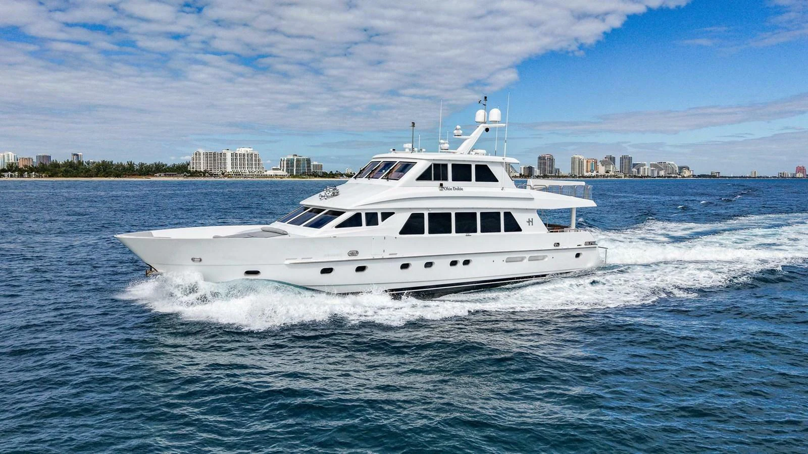 A sleek white yacht cruises through the open water with a cityscape in the background. aboard BOBBI TOO Yacht for Sale