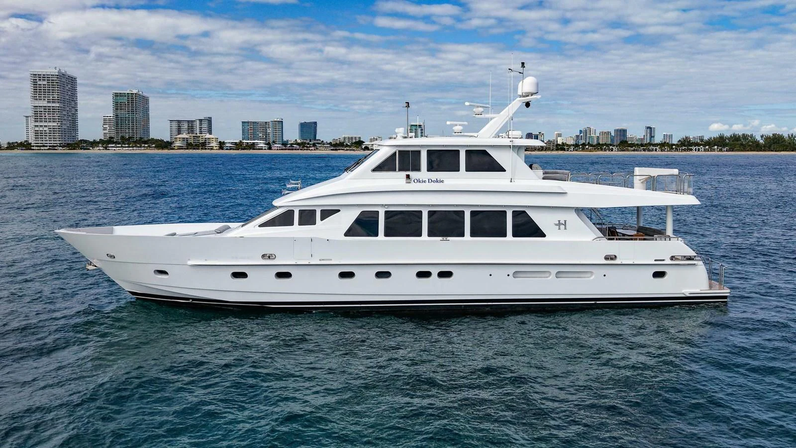 A sleek white yacht cruises gracefully on calm waters with a cityscape in the background. aboard BOBBI TOO Yacht for Sale
