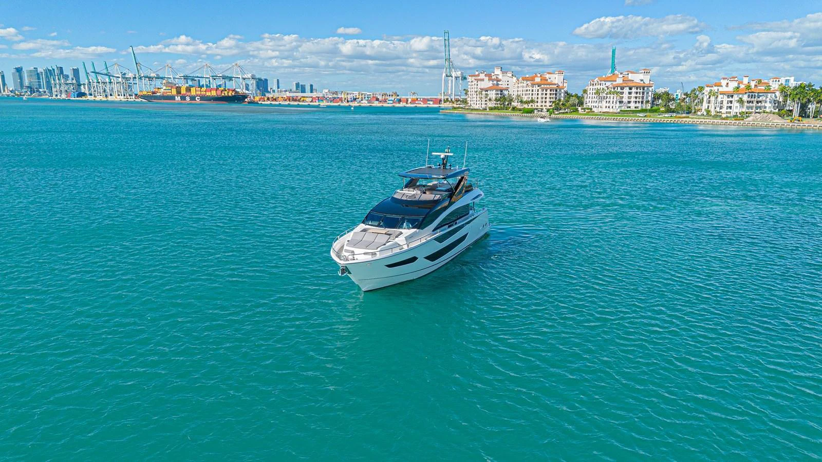 A sleek yacht glides through calm turquoise waters with a cityscape in the background. aboard HIDEOUT Yacht for Sale