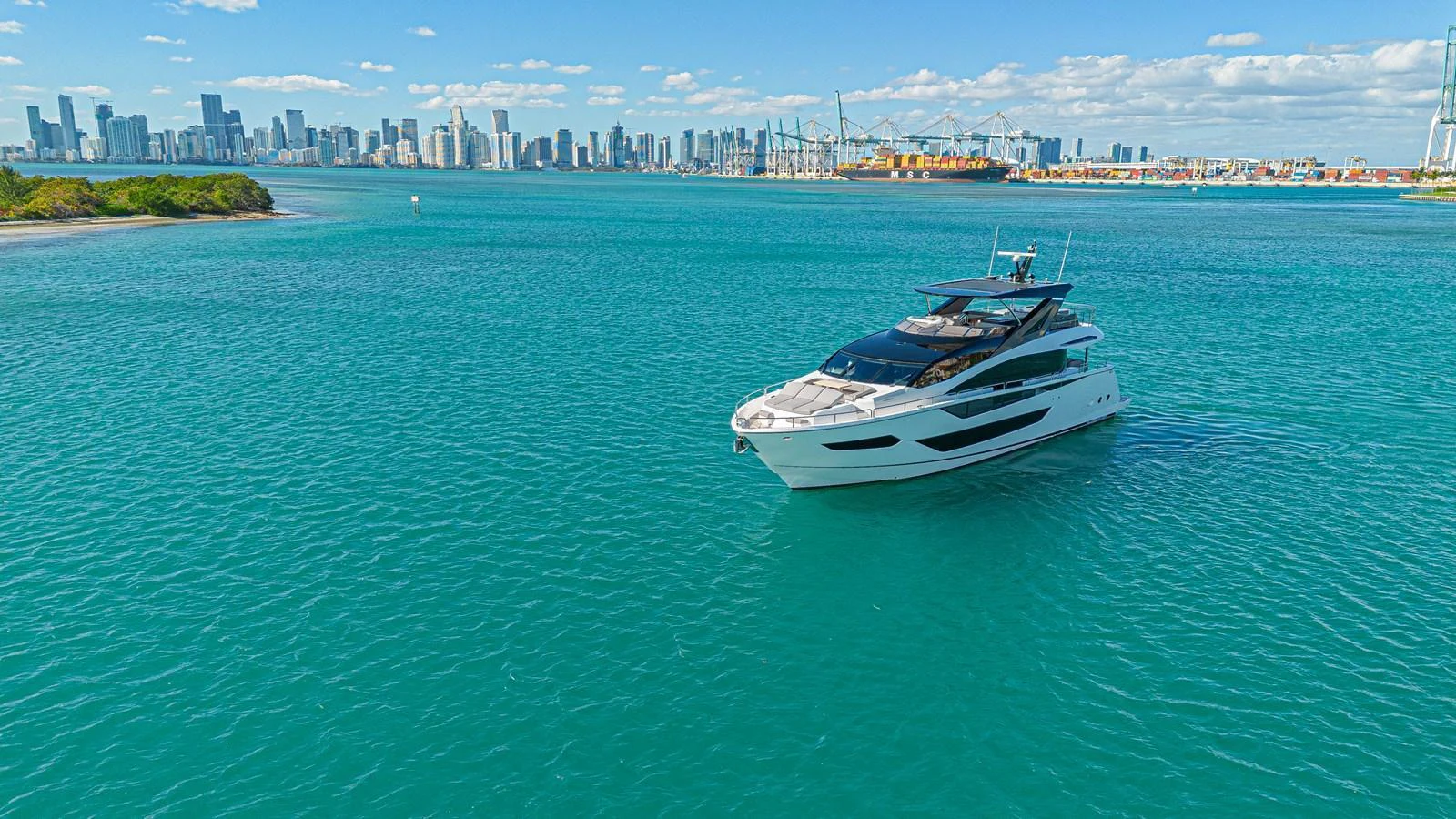 A sleek yacht cruises through turquoise waters with a city skyline in the background. aboard HIDEOUT Yacht for Sale