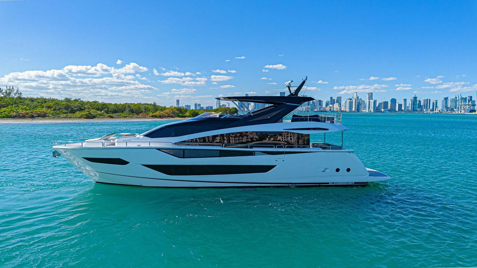 A sleek and modern yacht cruising in clear blue waters with a city skyline in the background. aboard HIDEOUT Yacht for Sale