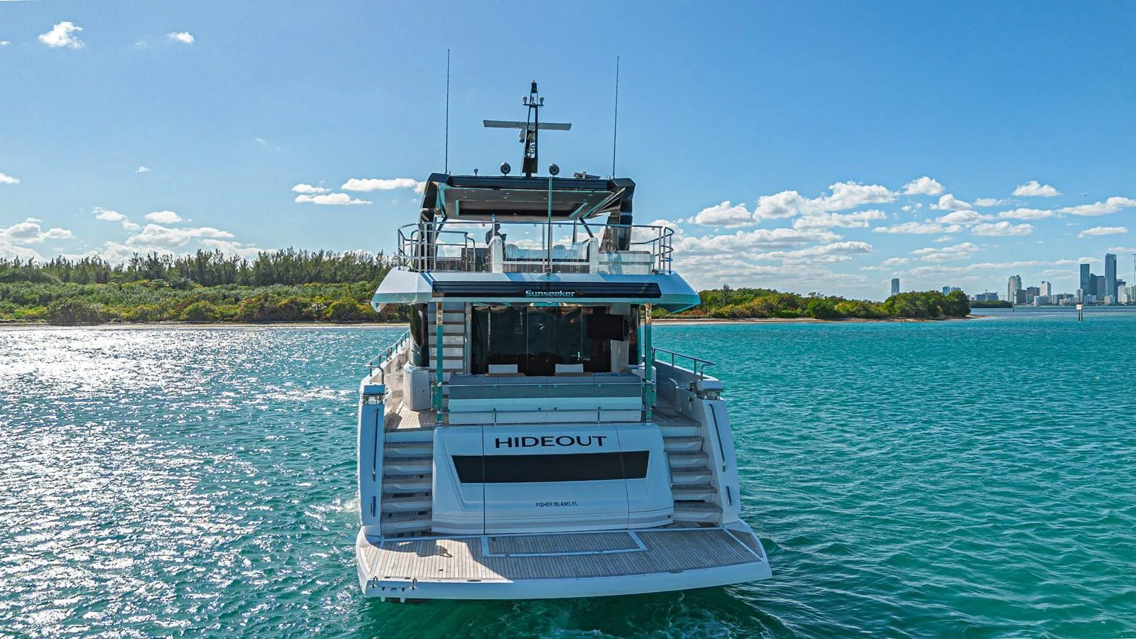 The yacht is seen from the rear, cruising in clear blue waters with a cityscape in the background. aboard HIDEOUT Yacht for Sale