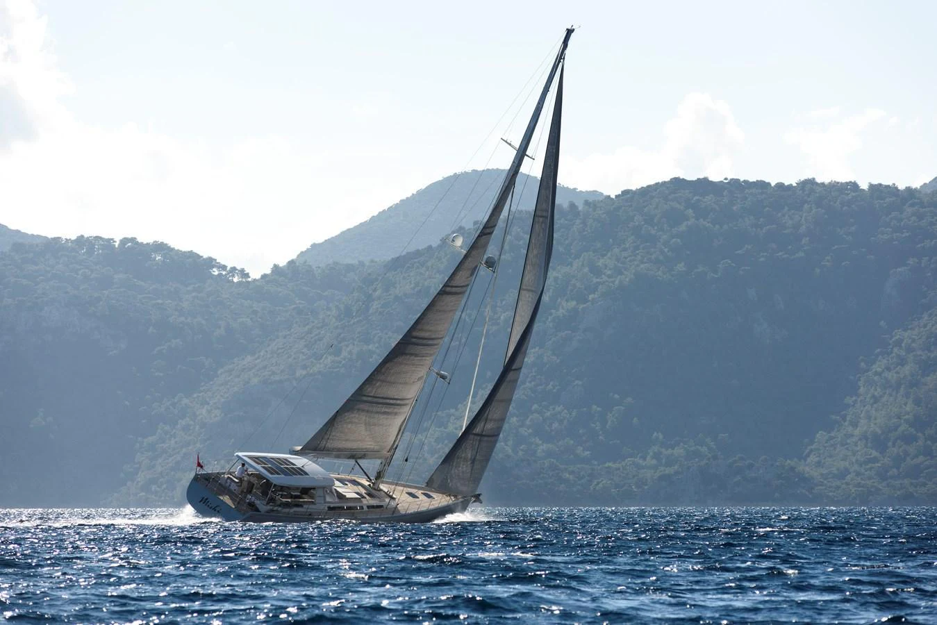 A sleek sailing yacht glides gracefully through the water against a backdrop of scenic hills. aboard MISHI Yacht for Sale