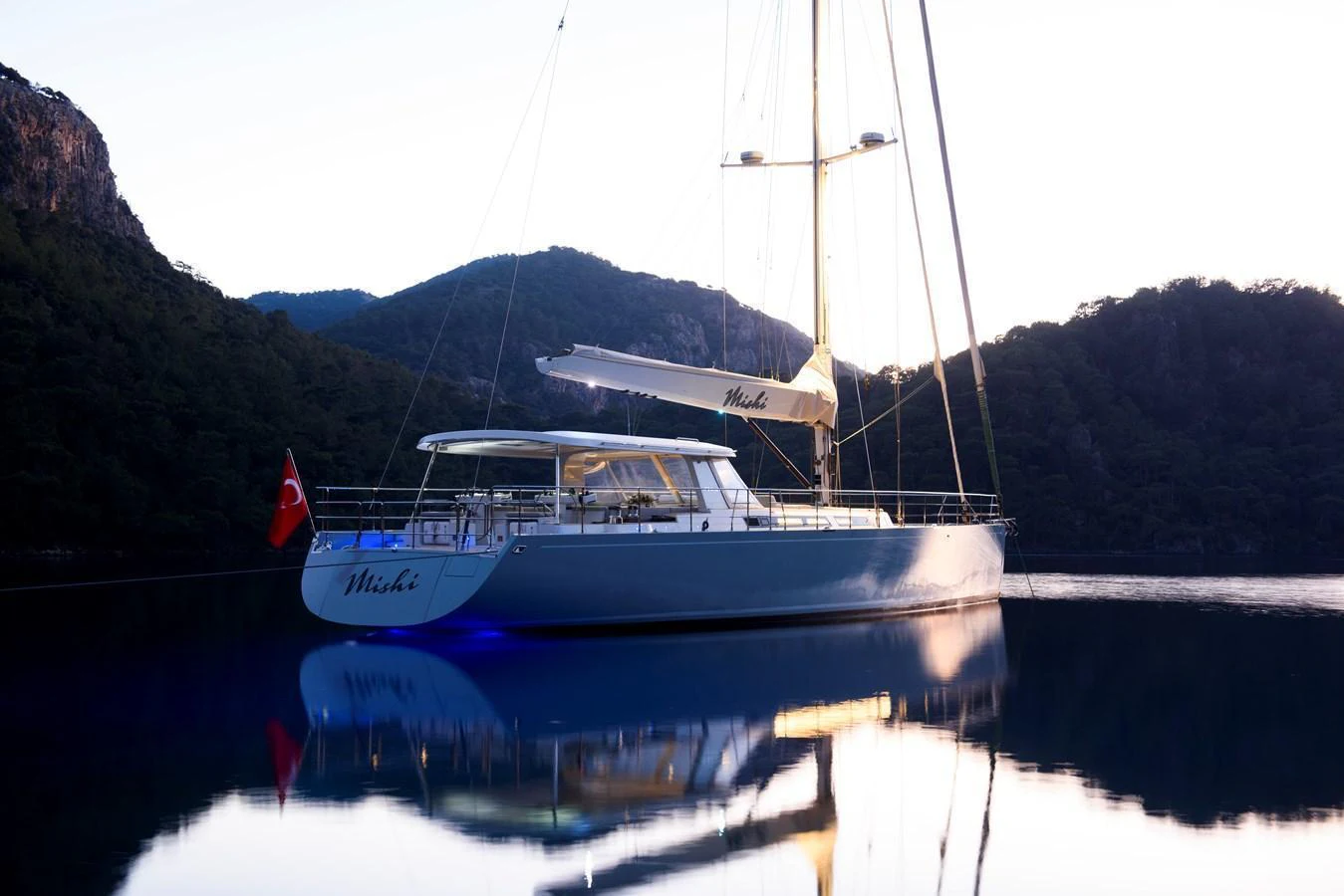 A sleek yacht is anchored in a serene, mountainous bay at dusk. aboard MISHI Yacht for Sale