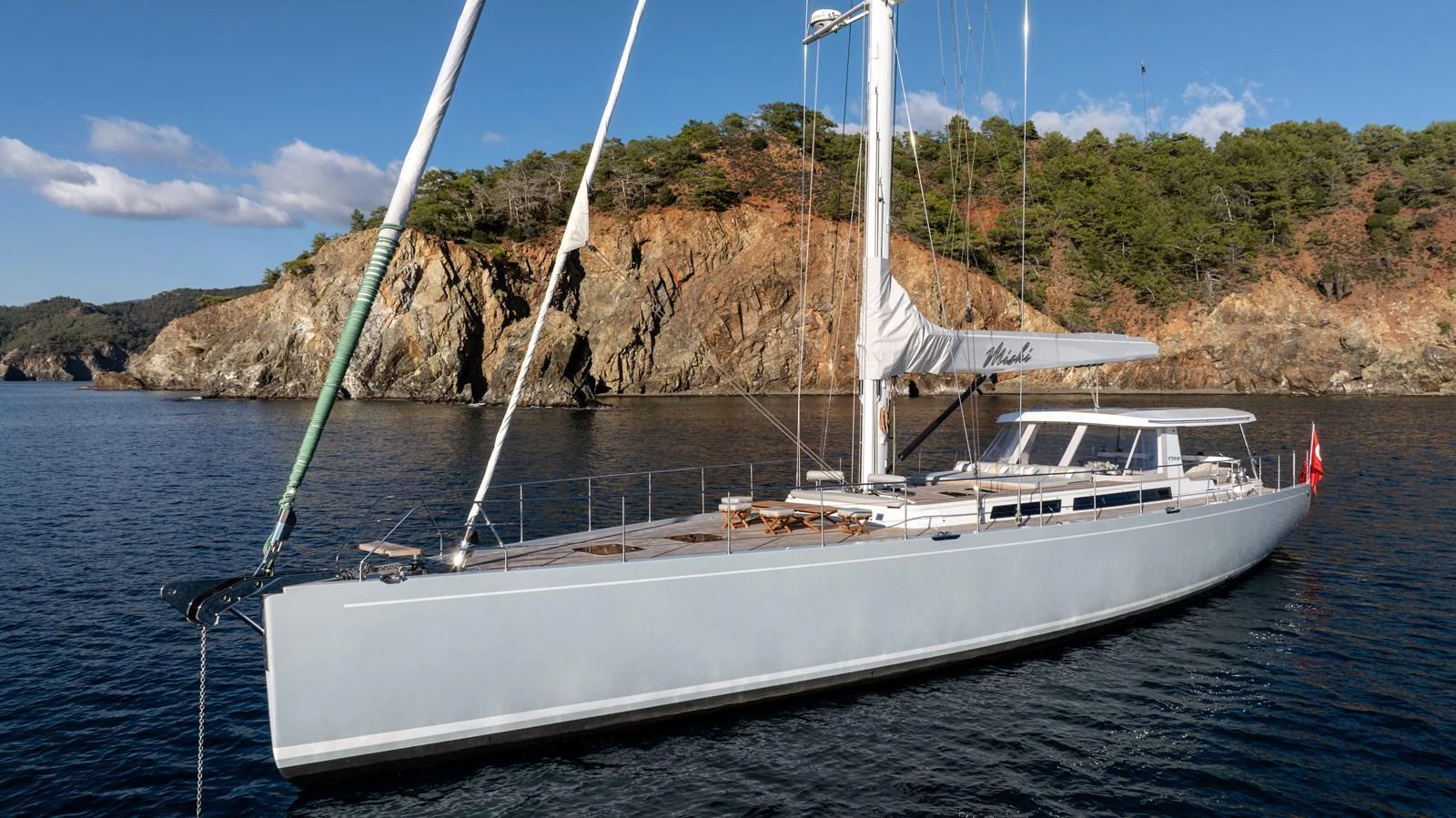 A sleek and modern sailing yacht glides gracefully on calm waters with a scenic rocky coastline in the background. aboard MISHI Yacht for Sale