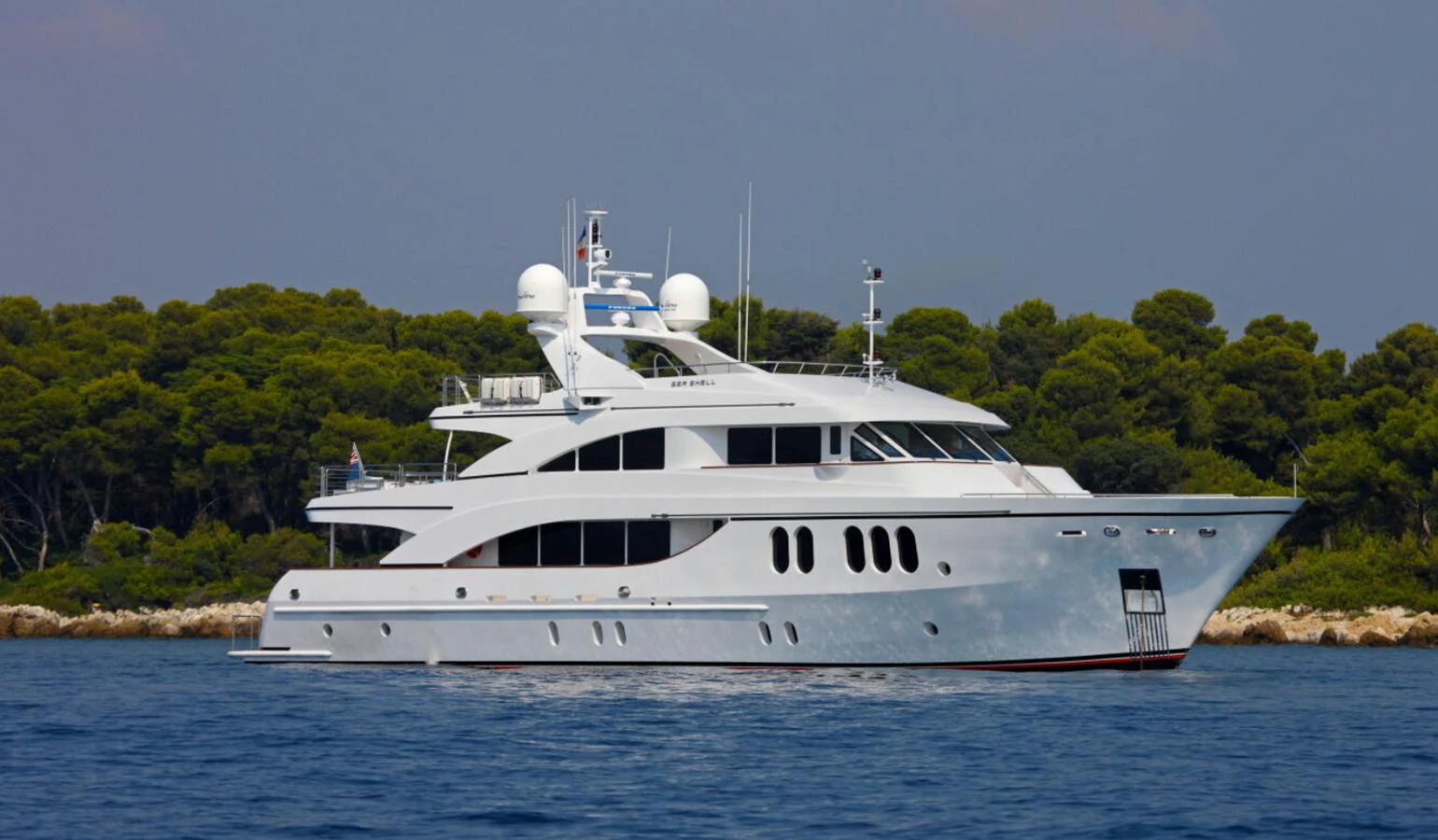 A sleek and elegant yacht is anchored near a lush, green shoreline. aboard SEA SHELL Yacht for Sale