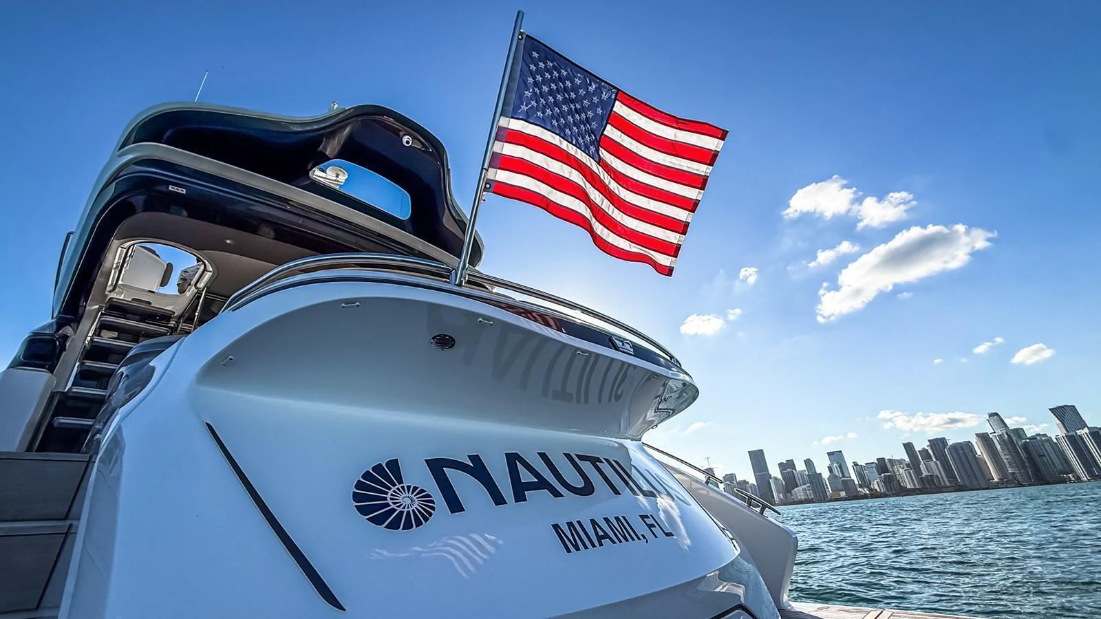 The stern of the yacht showcases its elegant design with a backdrop of a city skyline and a prominently displayed American flag. aboard NAUTILUS Yacht for Sale