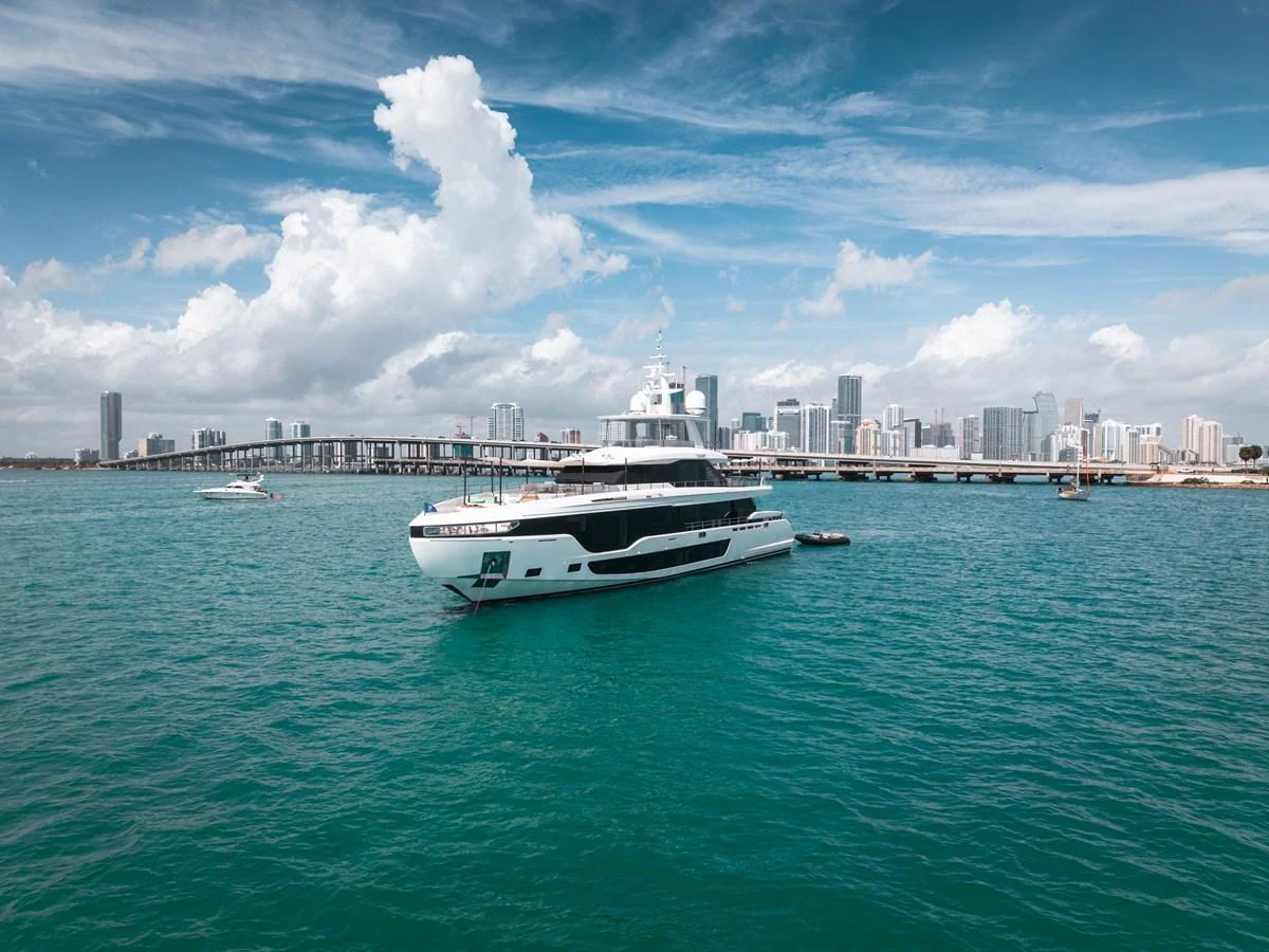 A sleek yacht cruises gracefully on turquoise waters with a cityscape in the background. aboard TYCOON Yacht for Sale