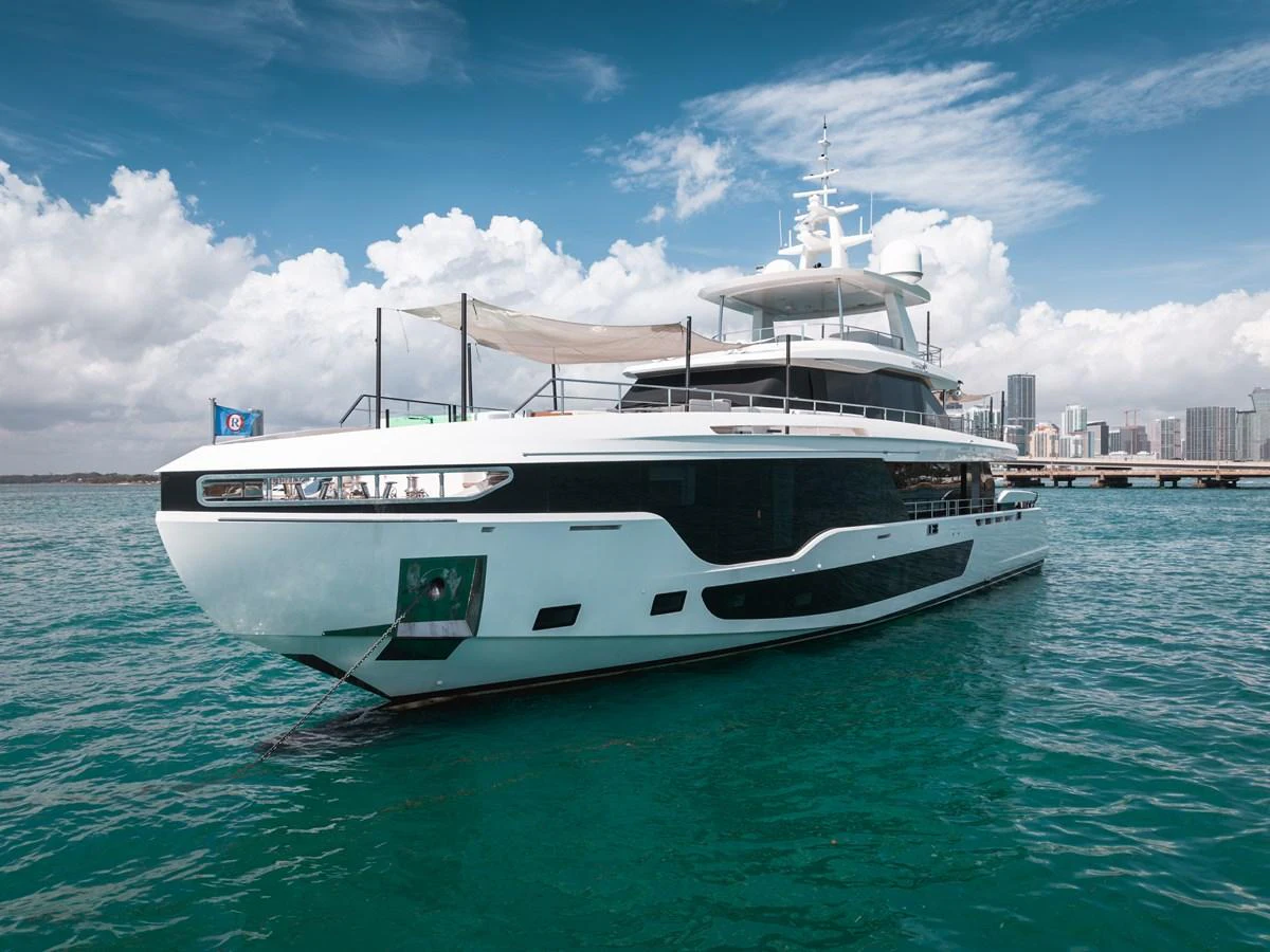 A sleek and modern yacht is anchored in clear turquoise waters with a city skyline in the background. aboard TYCOON Yacht for Sale