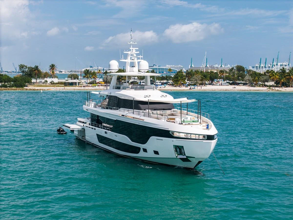 A luxurious yacht is anchored near a scenic coastline with clear blue waters and a backdrop of palm trees and cranes. aboard TYCOON Yacht for Sale