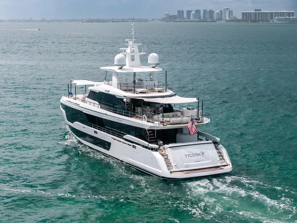 A luxurious yacht cruises through serene waters with a cityscape in the background. aboard TYCOON Yacht for Sale