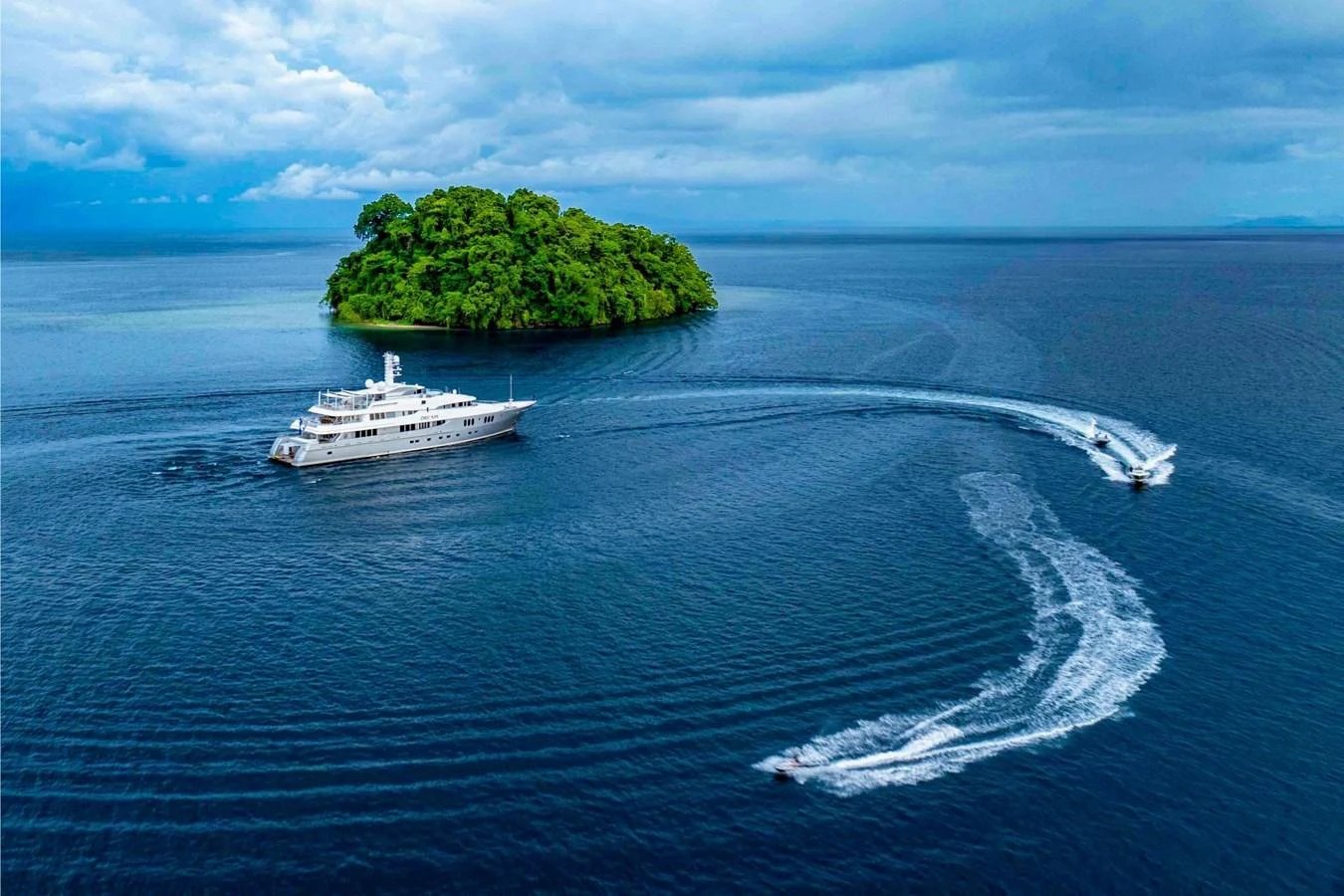 A luxurious yacht cruises near a lush, green island with a tender trailing behind. aboard DREAM Yacht for Sale