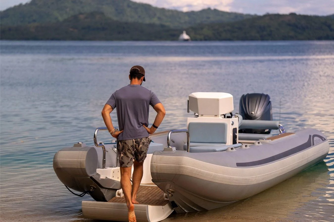 A man stands on the shore next to a sleek, modern tender boat with a scenic backdrop of calm waters and distant hills. aboard DREAM Yacht for Sale