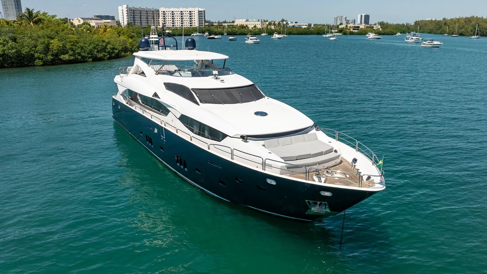 A sleek and modern yacht is anchored in a serene bay with lush greenery and buildings in the background. aboard BLUE Yacht for Sale