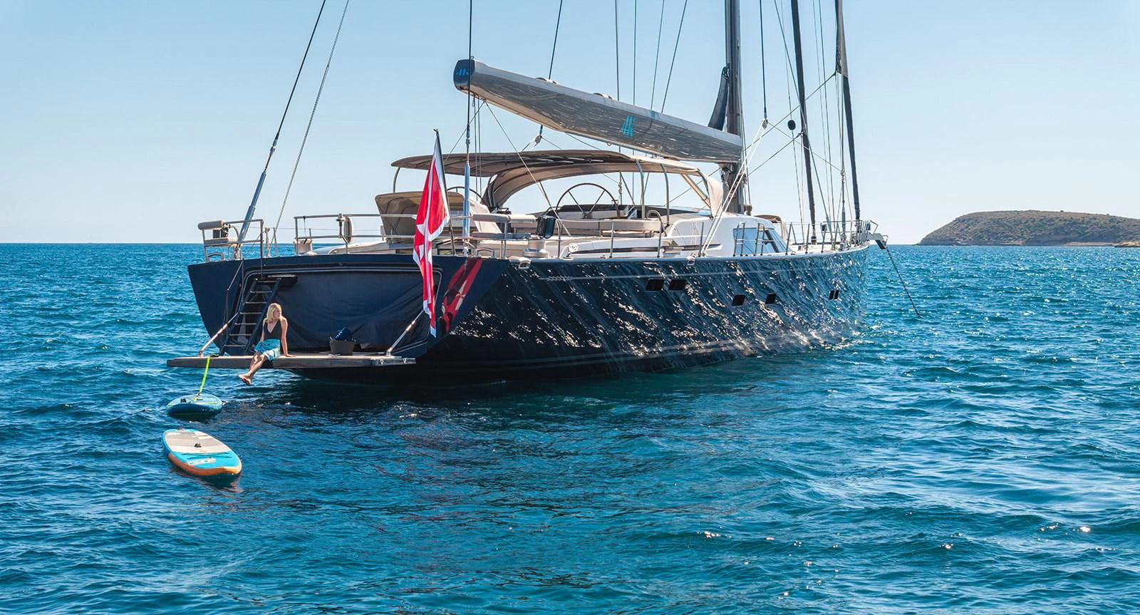 A sleek yacht anchored in calm blue waters with a paddleboard and a person enjoying the water at the stern. aboard 4K Yacht for Sale