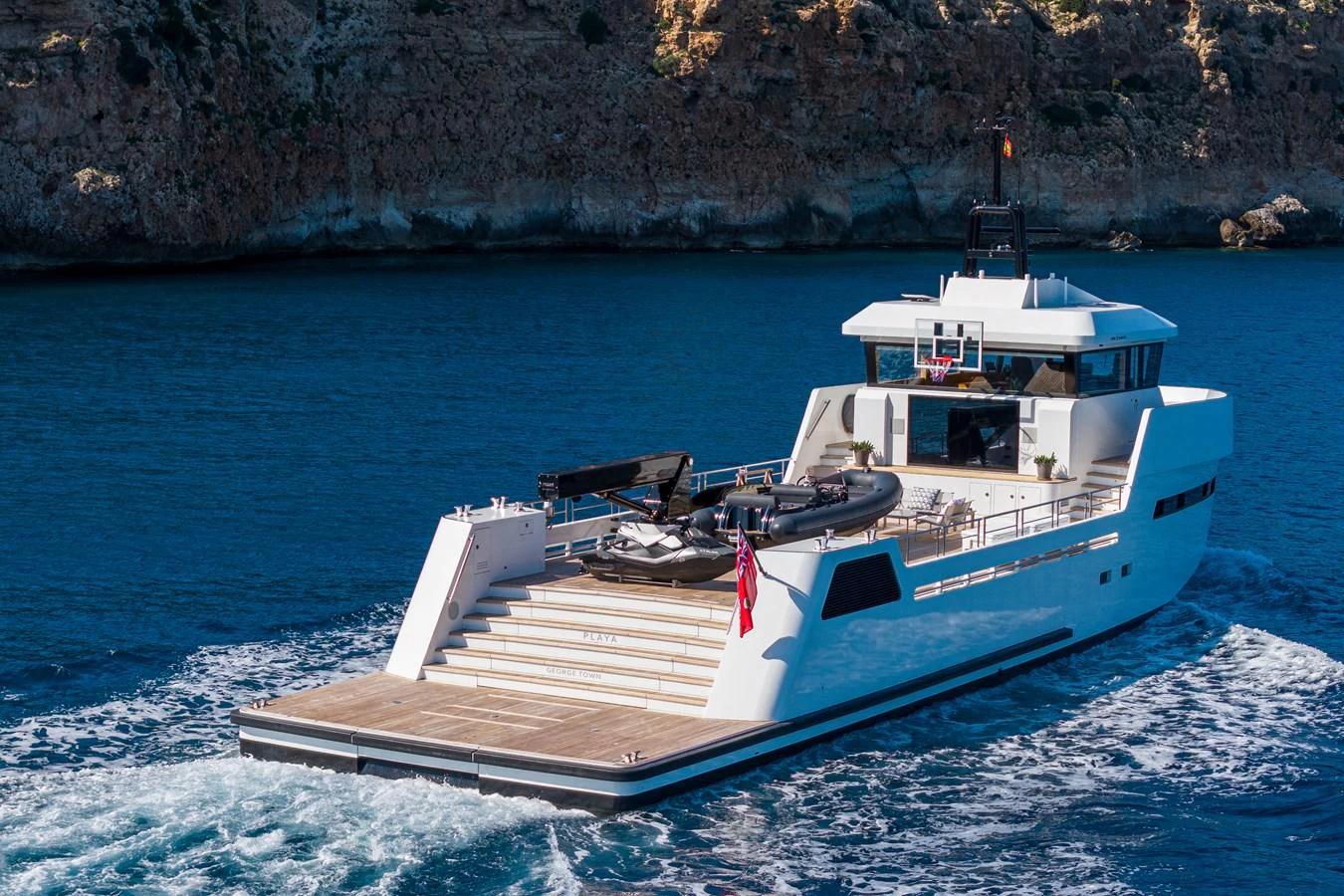 A sleek yacht cruising along a rocky coastline with a spacious aft deck and water toys. aboard PLAYA Yacht for Sale