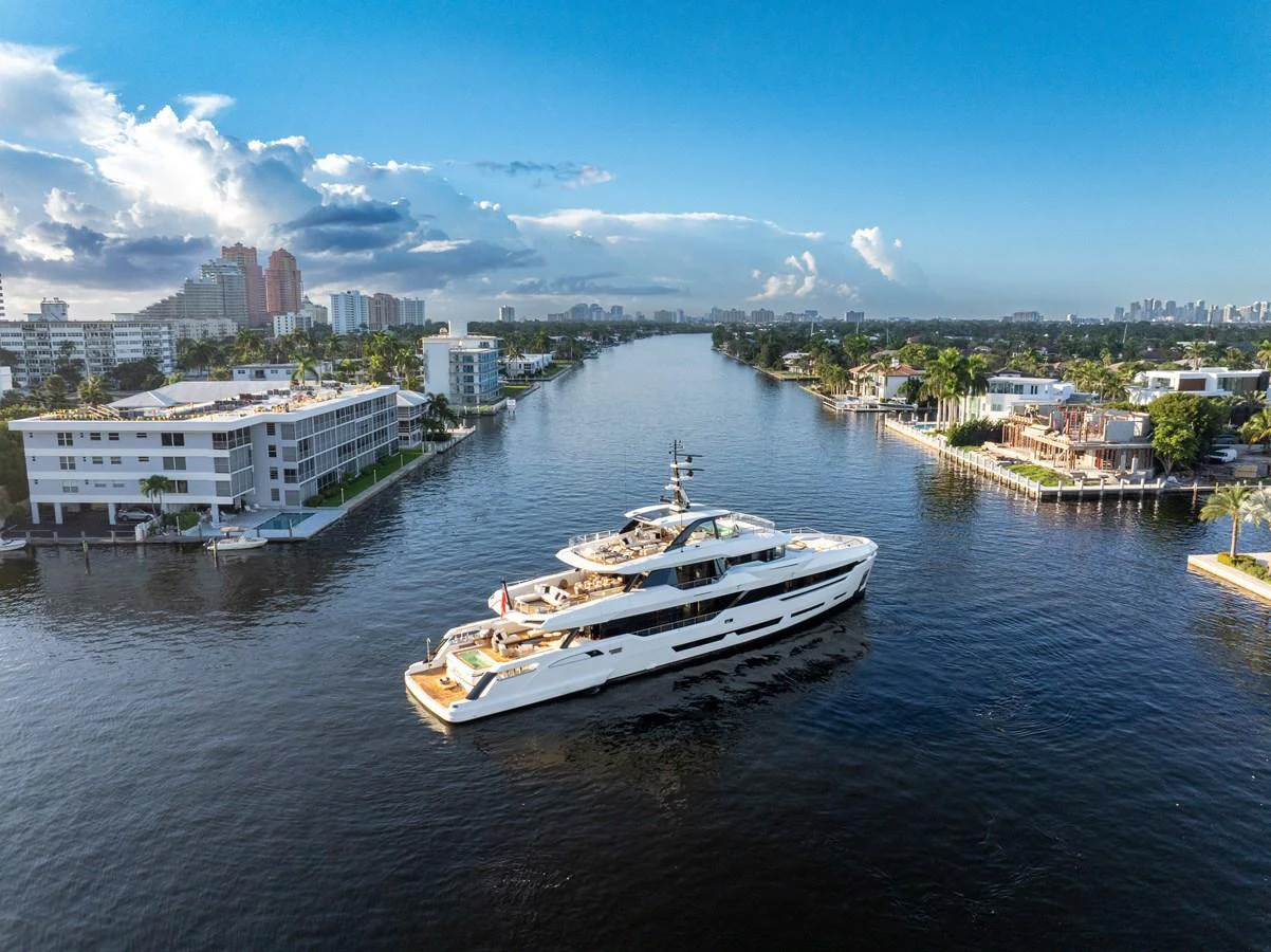 A luxurious yacht cruises through a scenic waterway flanked by modern buildings and lush greenery. aboard BAGLIETTO DOM 133 #5 Yacht for Sale