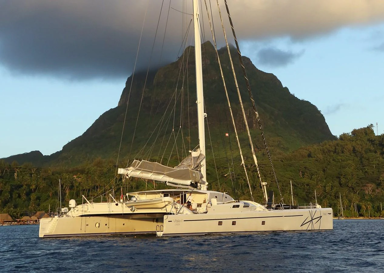 A sleek catamaran yacht sails gracefully against a backdrop of lush, mountainous terrain. aboard VITALIA II Yacht for Sale