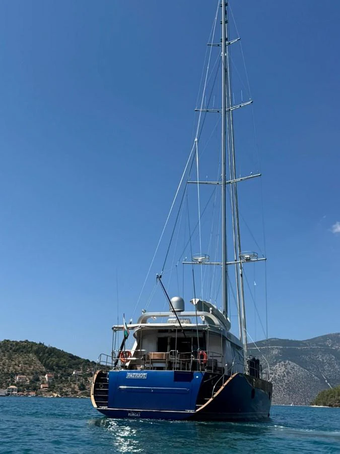 A stunning yacht is anchored in a serene bay with clear blue waters and a mountainous backdrop. aboard PATRIOT Yacht for Sale