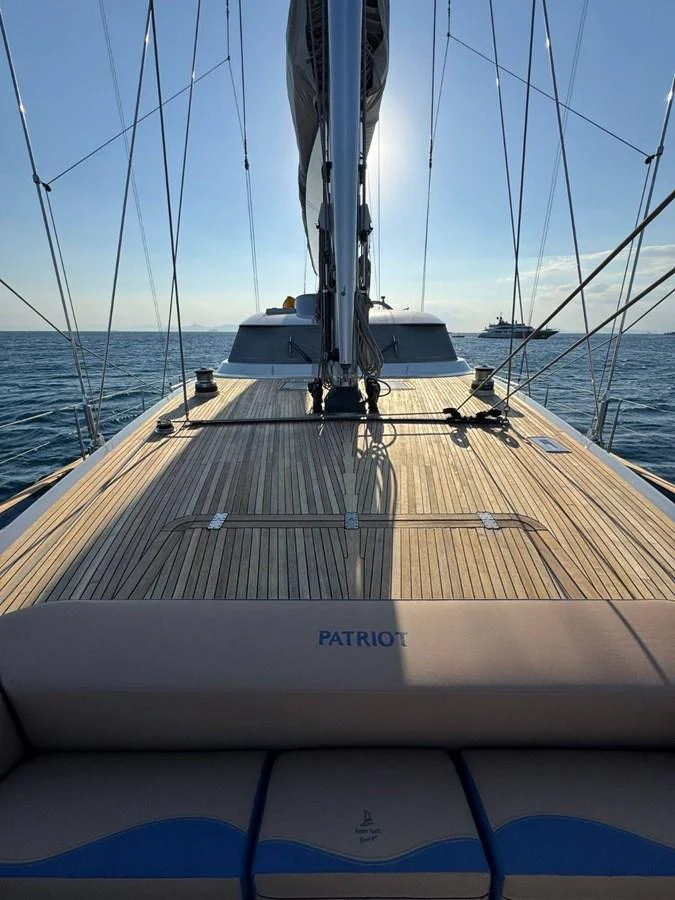 The yacht's deck showcases sleek wooden flooring with a clear view of the mast and sails, set against a serene ocean backdrop. aboard PATRIOT Yacht for Sale