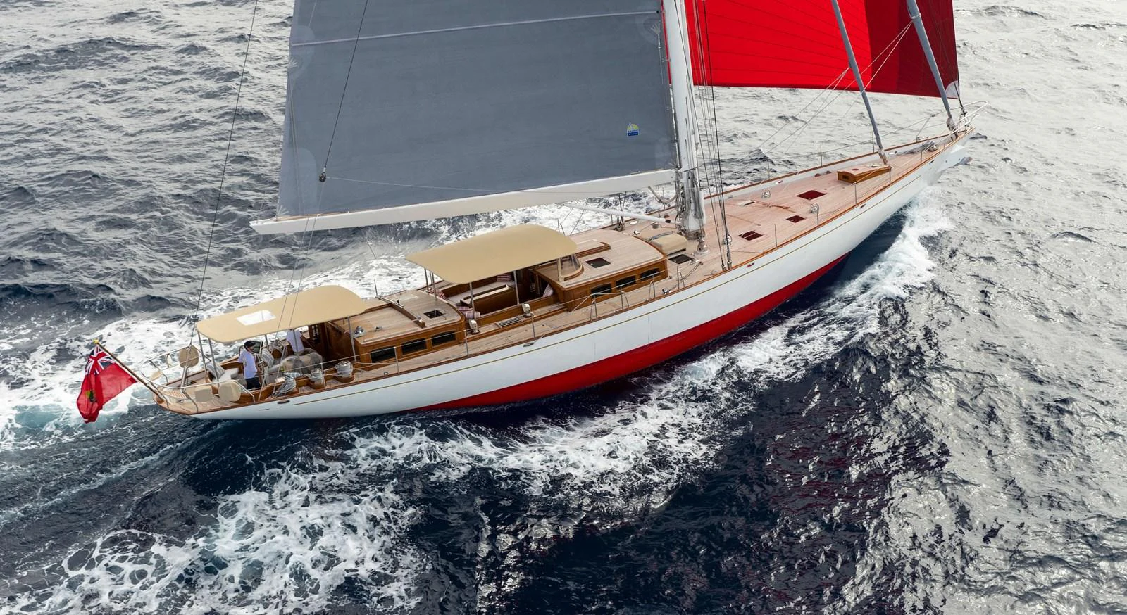 A sleek sailing yacht with red and grey sails glides through the waves. aboard ACADIA Yacht for Sale
