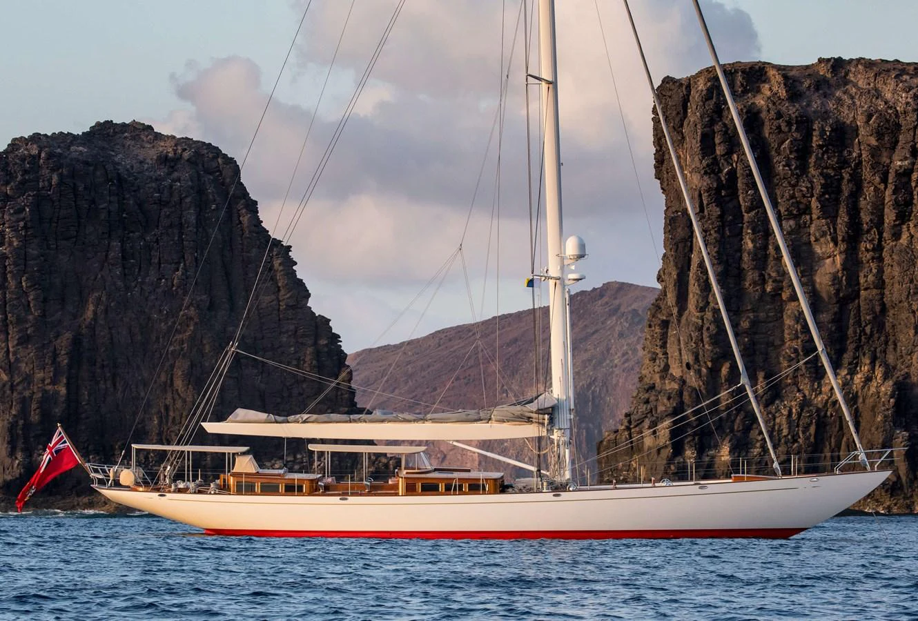 A sleek sailing yacht glides through calm waters with rocky cliffs in the background. aboard ACADIA Yacht for Sale