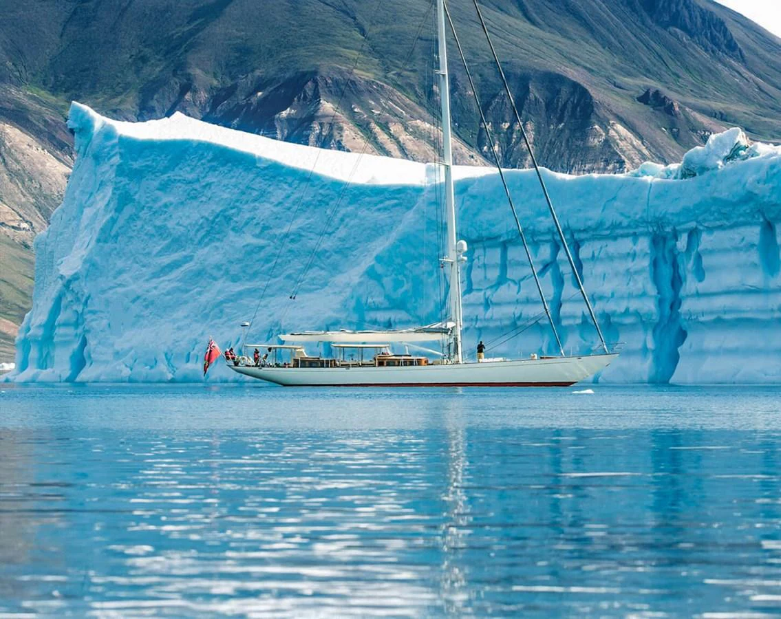 A sleek yacht sails gracefully past a majestic iceberg in serene, clear waters. aboard ACADIA Yacht for Sale