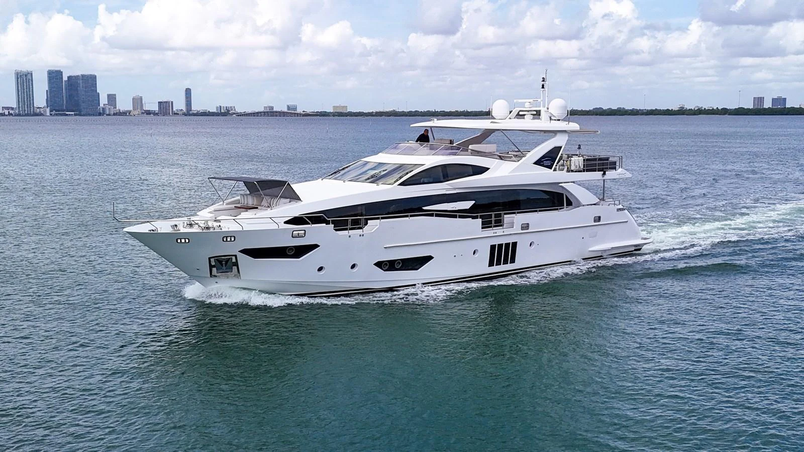 A sleek and modern yacht cruises through calm waters with a city skyline in the background. aboard 2017 AZIMUT 30 METRI Yacht for Sale