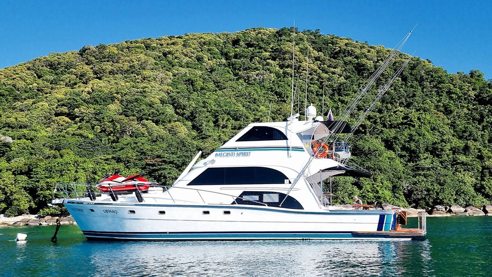 A sleek yacht is anchored in a serene bay with lush green hills in the background. aboard MEGISTI SPIRIT Yacht for Sale