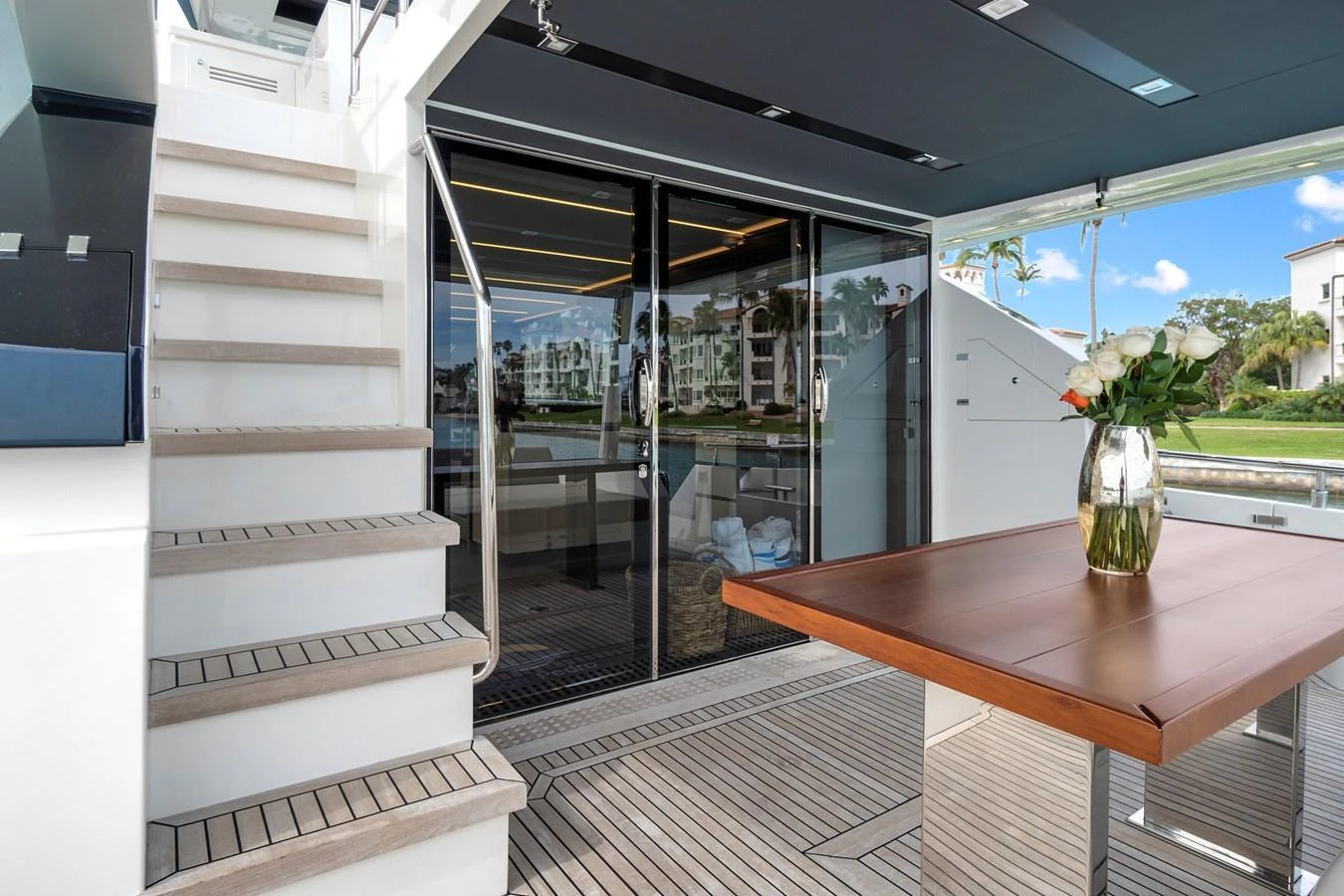 The aft deck features a stylish dining area with a wooden table and a vase of flowers, complemented by modern stairs leading to the upper deck. aboard JESSEA Yacht for Sale
