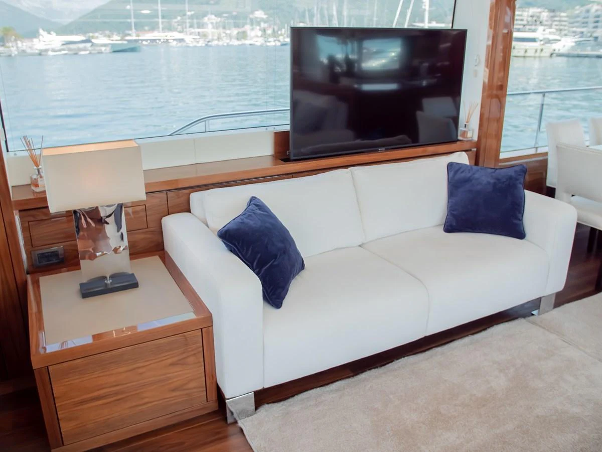 The salon features a comfortable white sofa with blue accent pillows, a wooden side table, and a flat-screen TV, all set against a backdrop of large windows offering a view of the marina. aboard SUNSEEKER 76Y Yacht for Sale