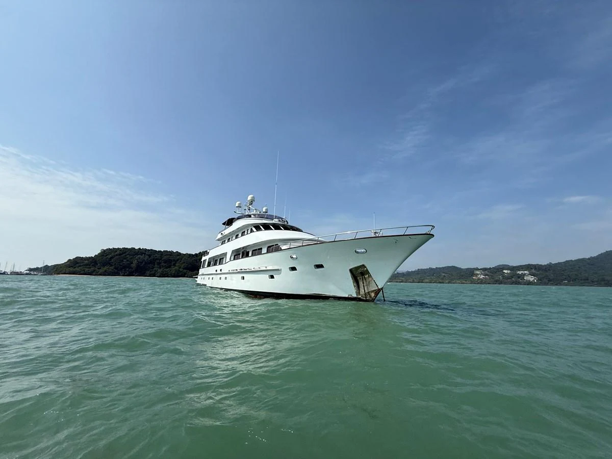 A luxurious yacht is anchored in calm, turquoise waters with a scenic backdrop of lush green hills under a clear blue sky. aboard LIGAYA Yacht for Sale