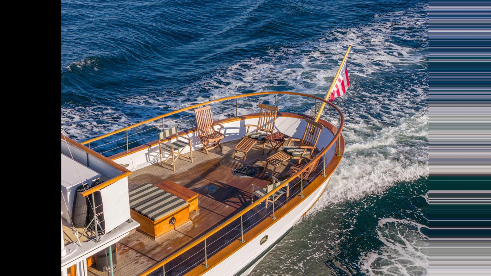 The yacht's aft deck features elegant wooden lounge chairs and a flag waving in the breeze as it cruises through the water. aboard BLUE PETER Yacht for Sale