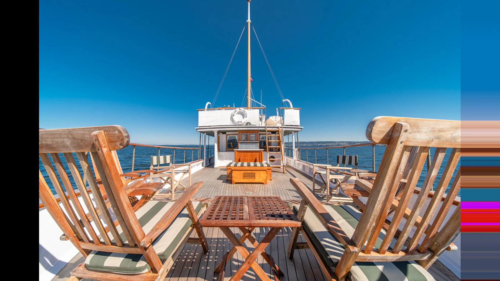 The yacht's aft deck features elegant wooden seating arrangements under a clear blue sky. aboard BLUE PETER Yacht for Sale