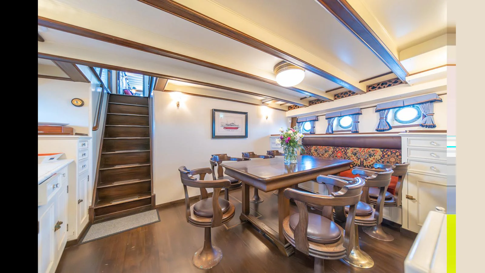 The yacht's dining area features a cozy and elegant setting with a wooden table and chairs, complemented by tasteful decor and natural light from portholes. aboard BLUE PETER Yacht for Sale