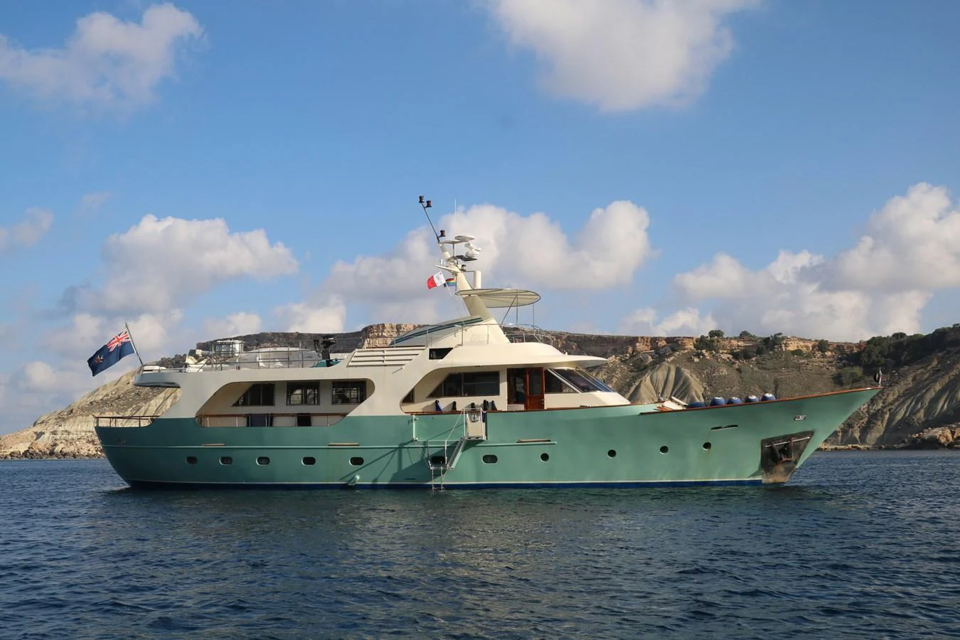 A classic yacht with a sleek design is anchored in calm waters against a scenic coastal backdrop. aboard KALANI Yacht for Sale
