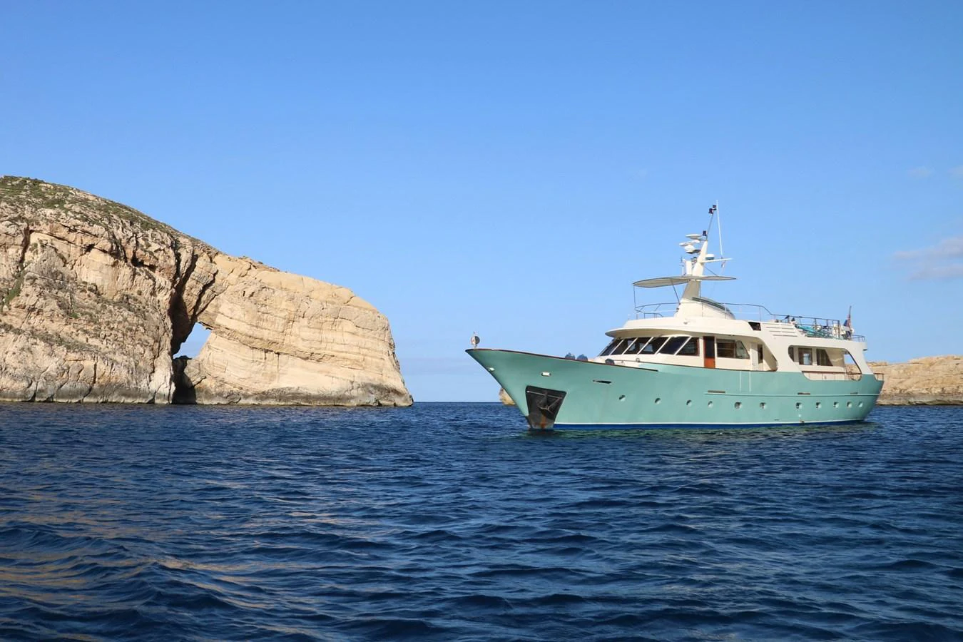A classic yacht cruises through serene blue waters near a rocky arch formation. aboard KALANI Yacht for Sale