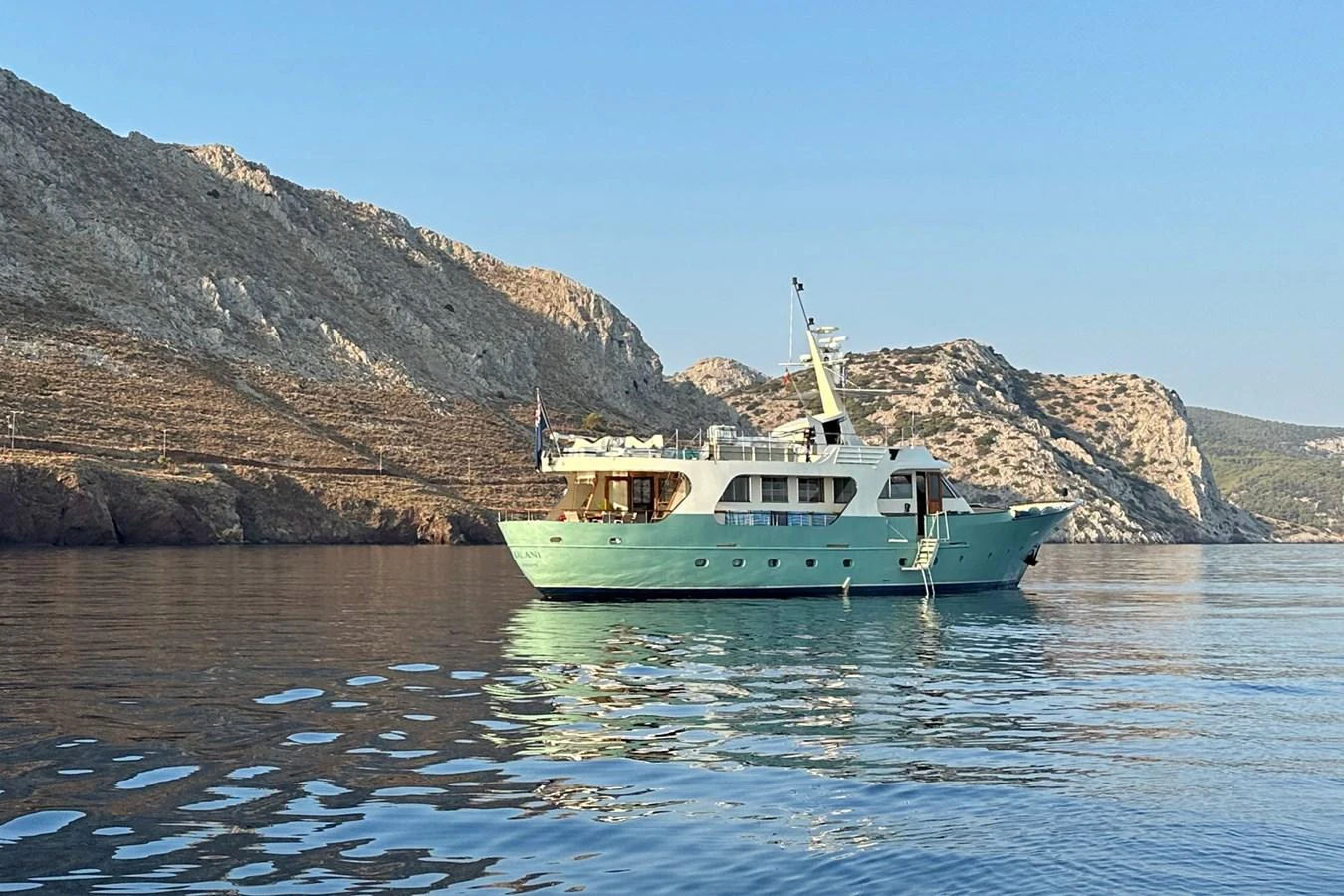 A classic yacht is anchored in a serene bay with rocky hills in the background. aboard KALANI Yacht for Sale
