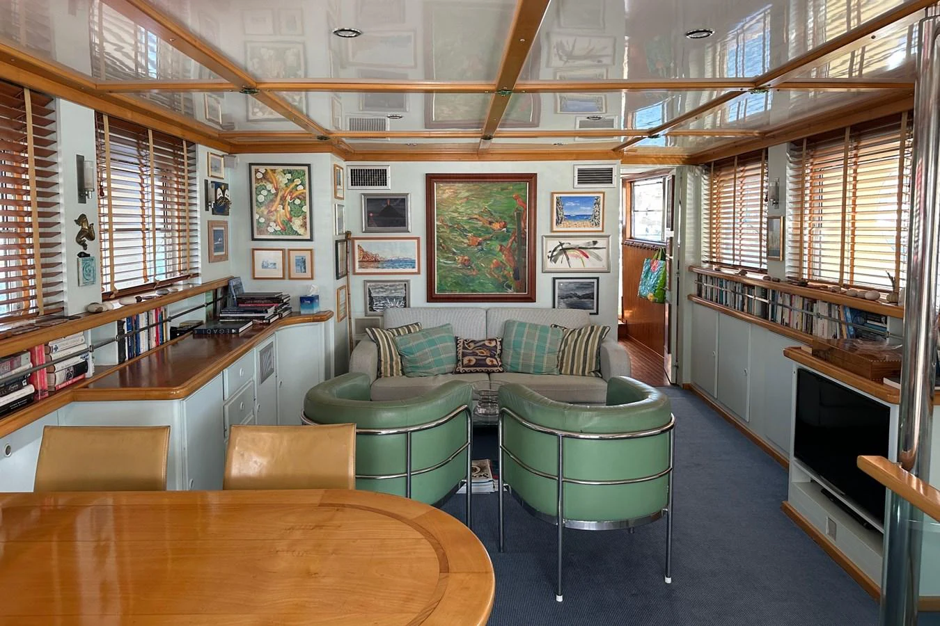 The yacht's interior features a cozy seating area with green leather chairs and a wooden dining table, surrounded by artwork and ample natural light. aboard KALANI Yacht for Sale