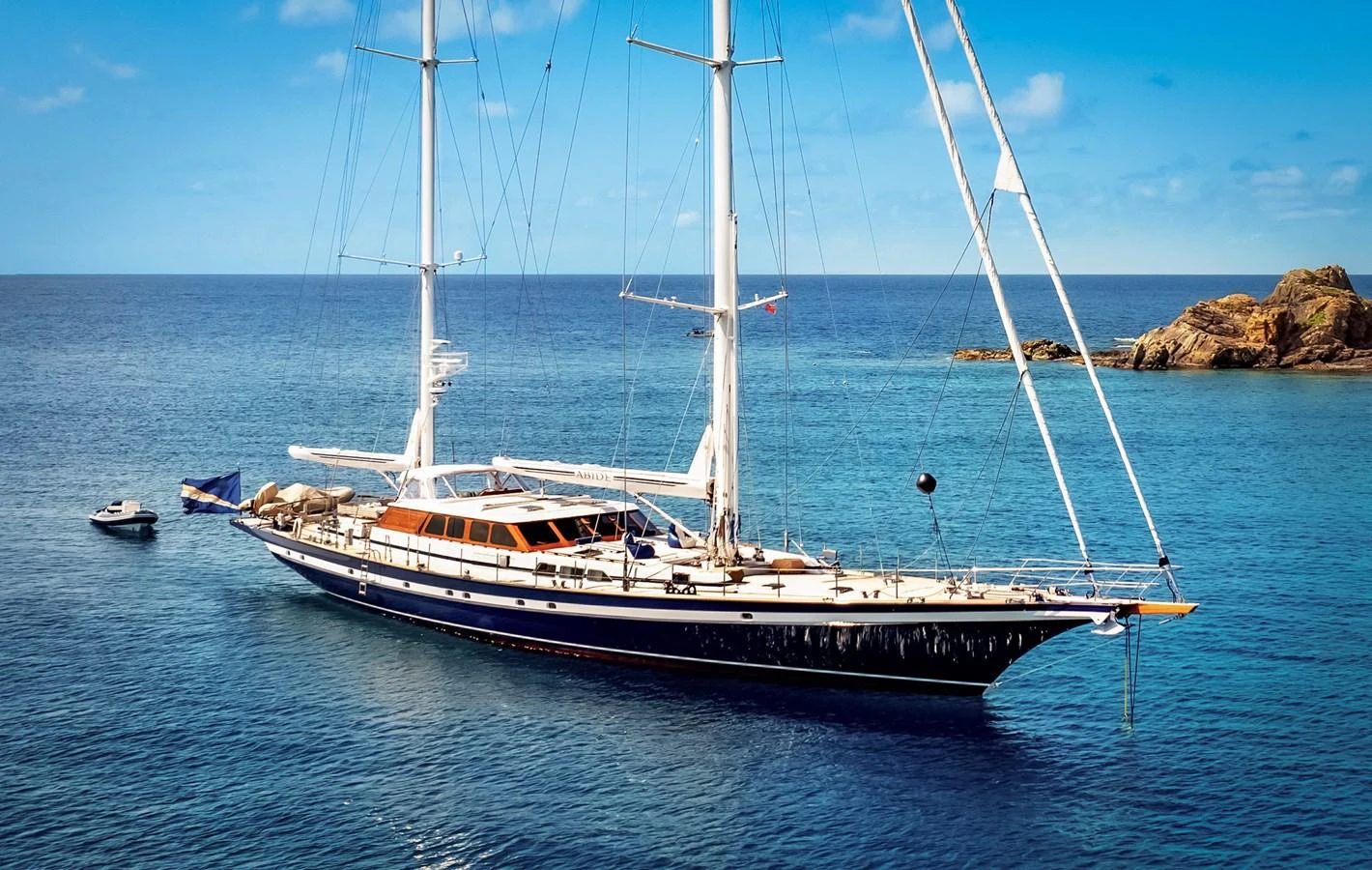 A stunning sailing yacht glides gracefully on calm blue waters near a rocky coastline. aboard ISLANDIA Yacht for Sale